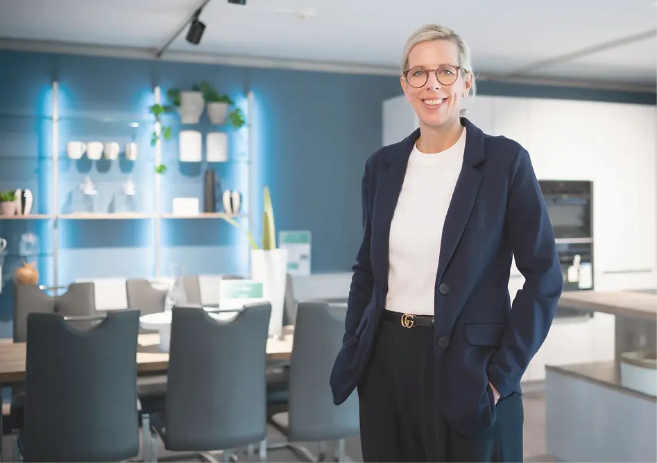 Frau steht im Küchenstudio vor Esstisch und Küchenmöbeln, im Hintergrund Regale mit Geschirr und Beleuchtung